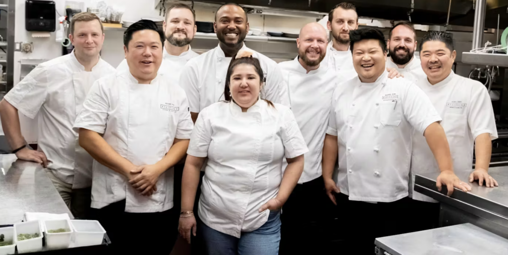 A large group of people in chef whites smile and pose in a kitchen.